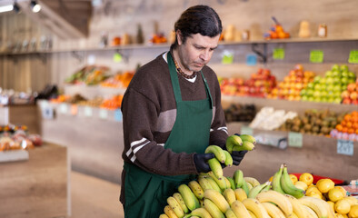 Man shop assistant puts ripe banana fruits in box on display case, arranges assortment in vegetable shop. Worker puts fruits in pile, in pyramid. Employee makes attractive display case