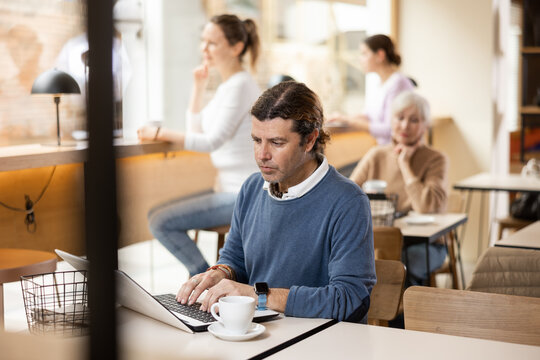 Positive European middle-aged man drinking and enjoying coffee while working on laptop in cafe. Freelance and remote work