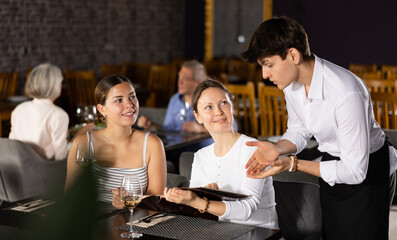 Two young girl friends resting, sitting in restaurant and ordering food, reading menu. Waiter guy tells young women visitors of cafe about dish from chef