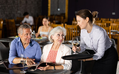 Senior male and female colleagues having nice time in sparsely populated cozy restaurant. Waitress answers questions of visitors, helps to choose diet dish for late dinner in menu