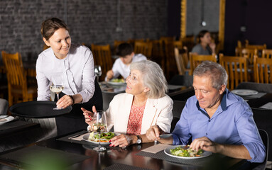 Adult woman waiter serving wine to couple of elderly man and woman in restaurant