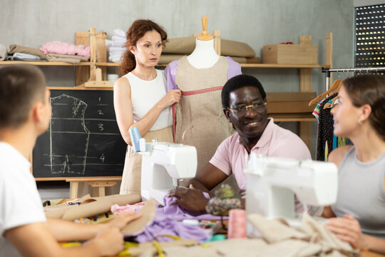 Experienced female instructor demonstrating garment measuring on mannequin while engaged adult students sewing and attentively listening to professional tips in hands-on fashion course..