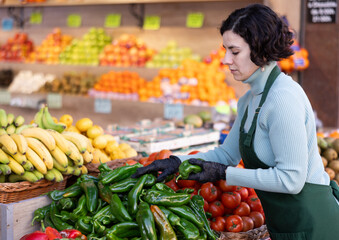 Adult woman seller in apron puts fresh bell peppers on display in vegetable shop