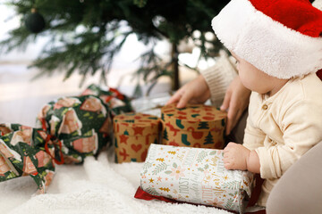 Happy cute baby in santa hat opening christmas present under xmas tree, first christmas. Merry Christmas and Happy Holidays! Joyful sweet child sitting with stylish christmas gifts