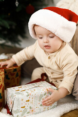 Cute baby opening stylish christmas gifts under decorated festive tree, close up. Merry Christmas and Happy Holidays ! Happy child in pajamas and santa hat playing with xmas presents
