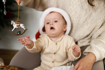 Happy cute baby in santa hat playing with christmas horse toy with mother under stylish xmas tree. Merry Christmas and Happy Holidays!