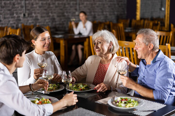 In cozy restaurant, young family and mature parents enjoy festive lunch, celebrating important event. Company chat and taste wine, enjoying pleasant atmosphere and good company.