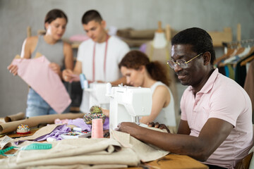 Engaged middle-aged man of African descent stitching fabric at machine during sewing class for adult beginners, while other participants preparing pattern pieces for assembly, working on fitting dress