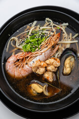 Seafood ramen with prawn, mussels and noodles in soy broth, black bowl on white background