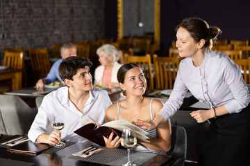 Newlyweds have lunch, spend time chatting in restaurant. Guy reads menu and makes order to waitress. Elderly couple is relaxing in cafe in background