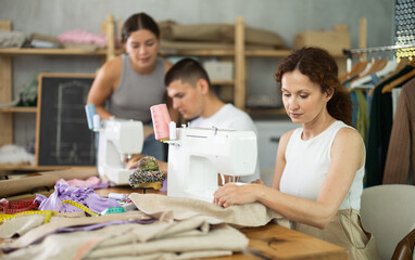 Glad middle-aged female student of dressmaking courses working with sewing machine in workshop