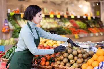 Adult woman seller in apron puts fresh pears on display in vegetable shop