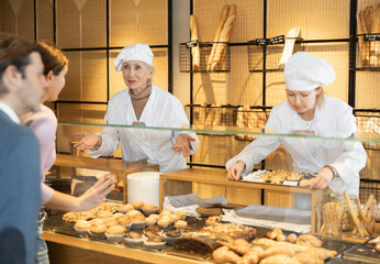 Portrait of two female bakery workers of different ages in uniform selling pastry to consumers in bakehouse