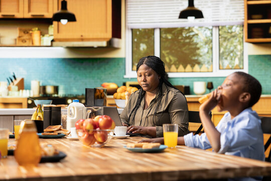 Busy mother ignoring children at breakfast table and focusing on her work, typing information on laptop and looking preoccupied. Distant working mom with a career and kids at home. - Powered by Adobe