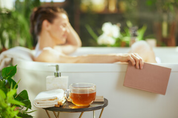 An adult woman, eyes closed, relaxes peacefully in a white bathtub, a book in hand. A tea mug & towels are arranged. Soft natural light illuminates green plants in this tranquil bathroom oasis.