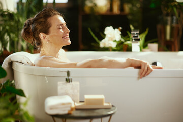 A middle-aged woman with a bun enjoys a peaceful bubble bath, smiling with closed eyes, purely relaxing. Soft lighting & blurred green plants frame scene, creating a tranquil sanctuary for self-care.
