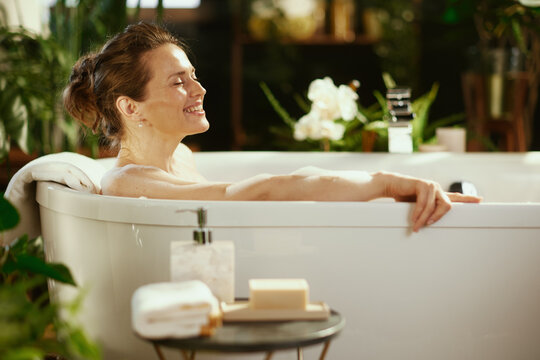 A middle-aged woman with a bun enjoys a peaceful bubble bath, smiling with closed eyes, purely relaxing. Soft lighting & blurred green plants frame scene, creating a tranquil sanctuary for self-care.