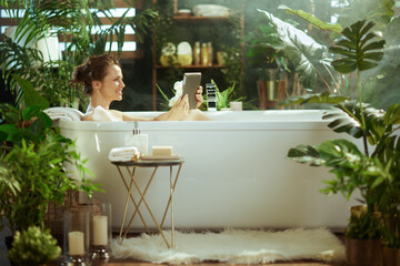 A smiling woman enjoys a luxurious bubble bath, holding a digital tablet in a bright, plant-filled bathroom. Steam & soft light enhance the serene, spa-like ambiance for relaxation & self-care.