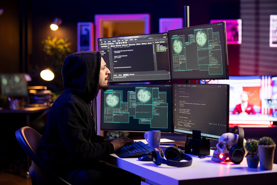 Hooded hacker in secret workspace bypassing firewalls, launching DDoS attacks on companies websites. Rogue technician at home working on virus scripts that can crash businesses servers