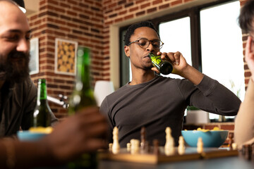 Black man with glasses, sips alcoholic beverage, contemplating next chess move. African american male player relaxes with beer, enjoying board game and snacks with friends in cozy living room.