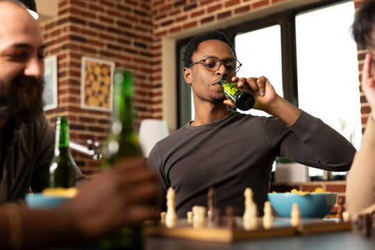 Black man with glasses, sips alcoholic beverage, contemplating next chess move. African american male player relaxes with beer, enjoying board game and snacks with friends in cozy living room.