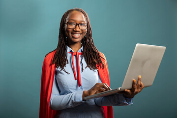 Portrait of black woman in blue shirt and red cape, standing confidently with laptop in hand. Female model embodying the image of corporate superhero, looking directly at camera.