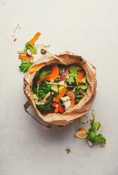 Fresh vegetable peelings and scraps in brown paper bag on light surface, highlighting food waste and its impact