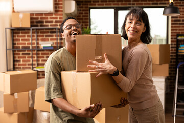 Portrait of multiethnic spouses standing in living room holding cardboard boxes. Happy African American man and Caucasian woman celebrate moving into new apartment, smiling joyfully at camera.