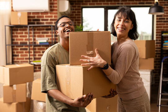 Portrait of multiethnic spouses standing in living room holding cardboard boxes. Happy African American man and Caucasian woman celebrate moving into new apartment, smiling joyfully at camera. - Powered by Adobe