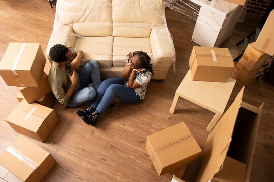 African american husband and wife rest indoors on floor near cardboard boxes and sofa, enjoying conversation and planning furniture. Black couple discussing cozy decorations in new home together.
