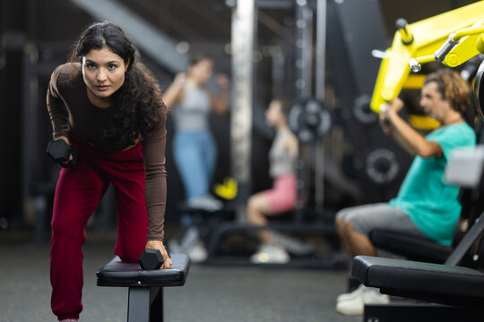 Latina woman standing near a bench in the gym and doing dumbbell rows for back muscles. Motivated woman doing upper body workout for relief arms and back