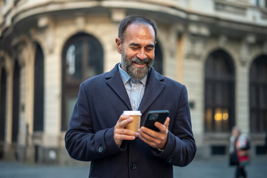 Mature man smiling using smartphone drinking coffee in city - Powered by Adobe