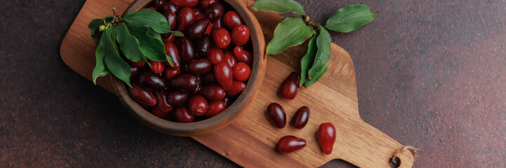 Close-up horizontal shot of cornelian cherries in a wooden bowl with green leaves on a rustic cutting board. Seasonal organic fruit for healthy lifestyle and wellness.