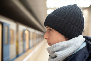 Profile of a teenage boy in a black hat, against the background of a train and a subway platform.