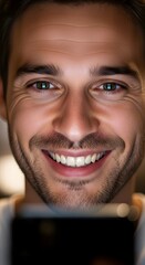 A close-up of a smiling young man with clear skin, bright eyes, and a friendly expression, looking at a smartphone screen with a joyful demeanor