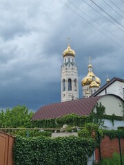 The golden domes and bell tower of an Orthodox church rising above a residential building and ivy-covered fence, set against a dramatic, stormy dark sky.