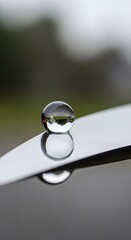A clear glass marble resting on a smooth, reflective surface outdoors with a blurred natural background under an overcast sky