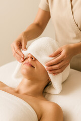 Vertical spa scene showing a therapist placing a warm, soft towel over a woman’s eyes as she relaxes on a treatment bed, capturing a soothing, restorative moment ideal for wellness centers, spa ads