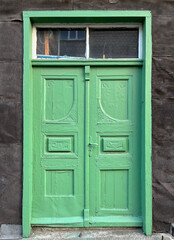 Vintage green double wooden door with decorative panels and a rectangular transom window, set in a dark textured wall, showing weathered paint and classic architectural details.