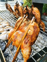 The image shows chicken pieces, including a whole leg and drumstick, being grilled over hot charcoal. The chicken is skewered with bamboo sticks and has a golden brown, slightly charred appearance.