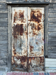 Close-up of a heavily rusted metal double door with peeling paint and corroded surface, set in a weathered wooden wall with horizontal planks and faded gray tones.