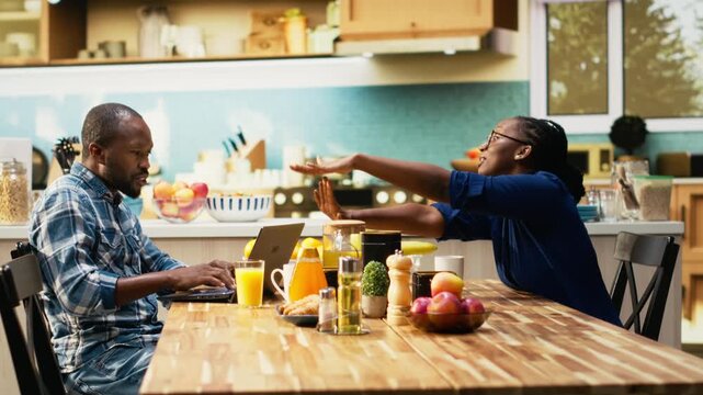African american man working too hard and woman asking for a break, showing him the timeout sign and closing his laptop. Workaholic boyfriend ignoring his girlfriend during breakfast. Camera B.
