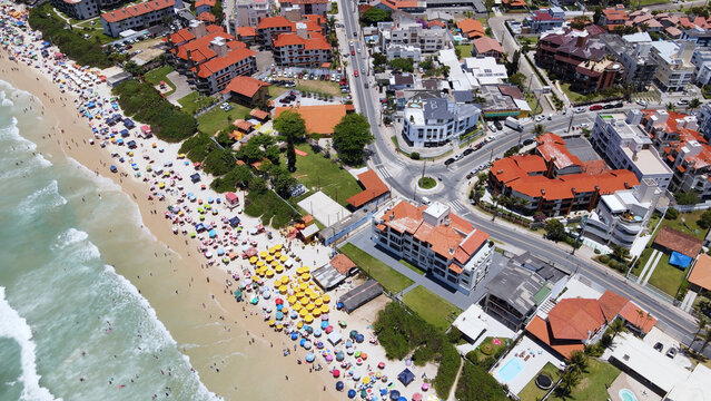Aerial view of the urban area of Ingleses beach in Florianópolis, Santa Catarina, showing streets and avenues near the sea with the sand crowded with people and umbrellas. - Powered by Adobe
