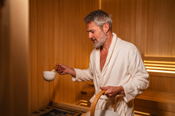 Mature man in bathrobe pouring water in sauna
