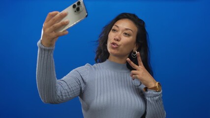 Woman taking selfie with smartphone, smiling and flashing peace sign while holding phone at arm's length in a blue studio; joy social connection.