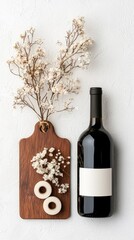 A dark bottle of wine with a blank label sits next to a wooden cutting board adorned with dried flowers and two small white rolls.
