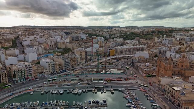 Aerial timelapse over Msida, Malta showing ongoing flyover construction beside the marina and St. Joseph Church, with urban traffic and dramatic clouds moving across the skyline.