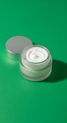 Open jar of creamy white moisturizer with silver lid placed on vibrant green background for skincare or beauty product display