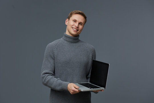 smiling young caucasian man holding laptop wearing grey knitted turtleneck look camera isolated on plain gray background digital nomad technology concept
