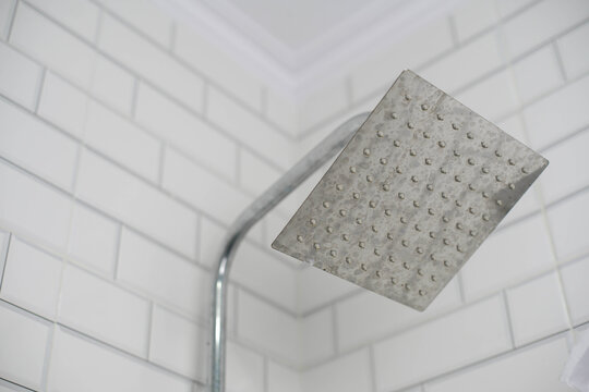 Close-up of a square rain shower head with visible hard water spots against clean white subway tiles, highlighting bathroom maintenance challenges.